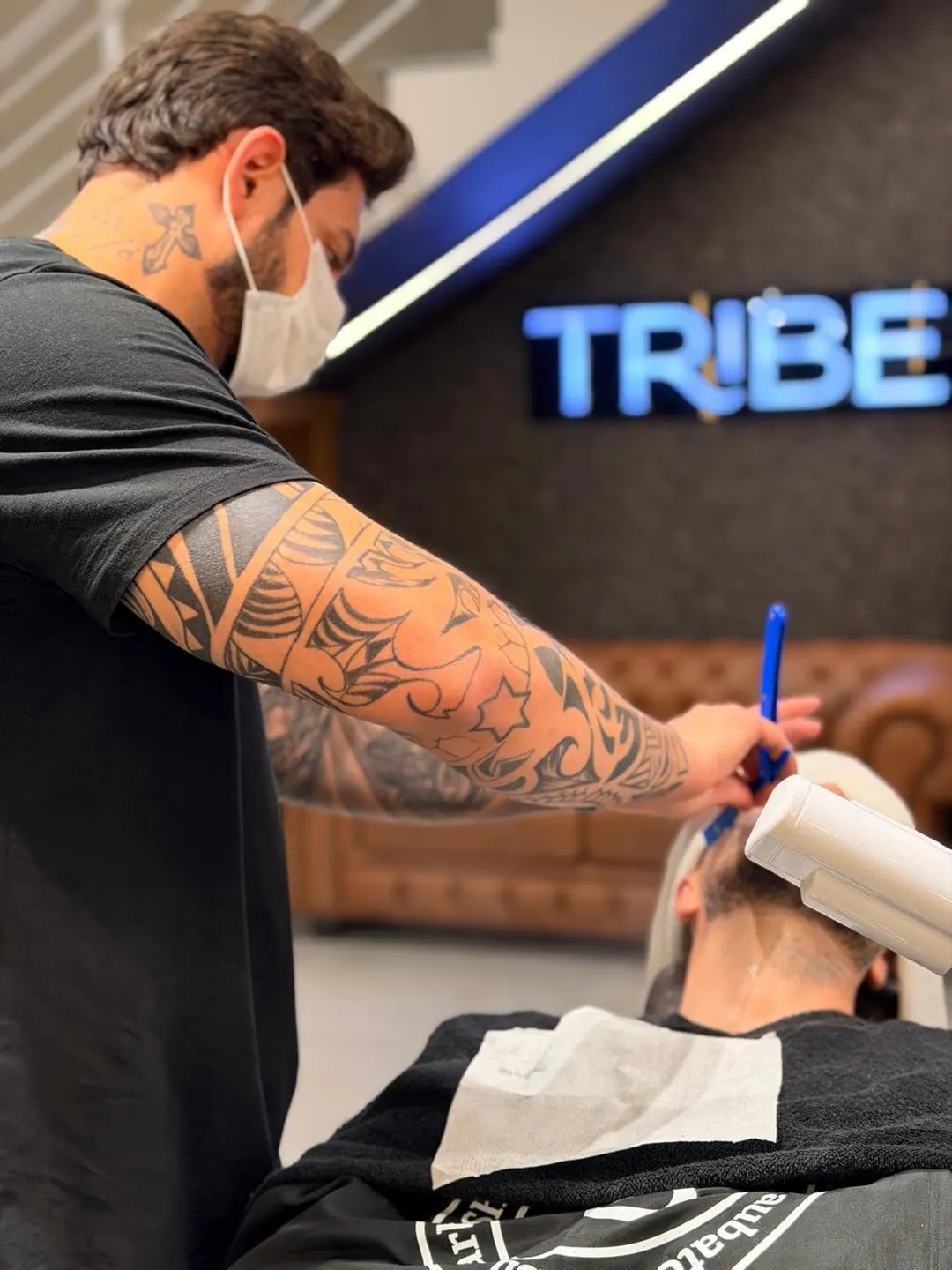Barbeiro da Tribe cortando cabelo sob o letreiro neon TRIBE, na barbearia onde acontecem as aulas